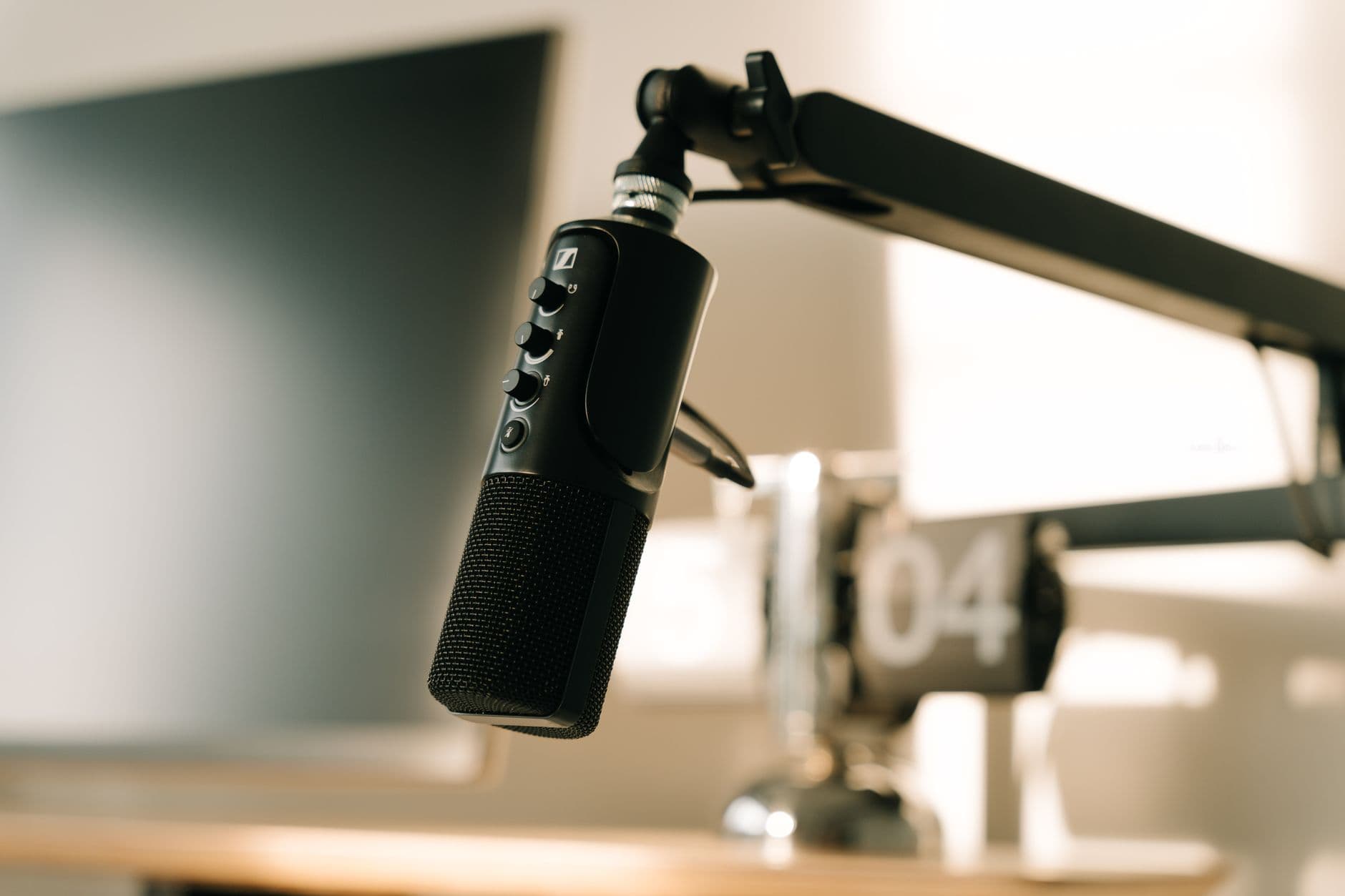 Close-up of a professional microphone on a desk with audio editing software on a monitor in the background.
