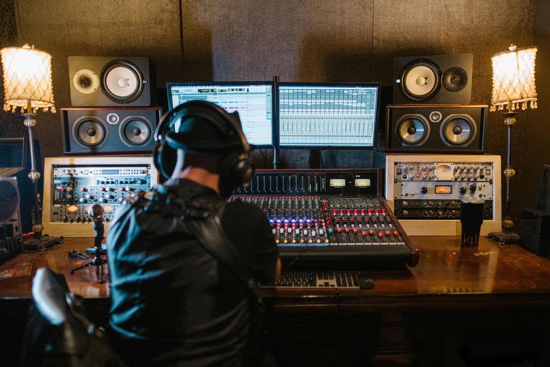 Audio engineer's back view at a mixing console with dual monitors displaying editing software.