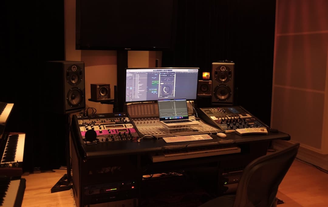 A dark studio desk with multiple monitors displaying audio editing software and a laptop for faceless YouTube channel operations.