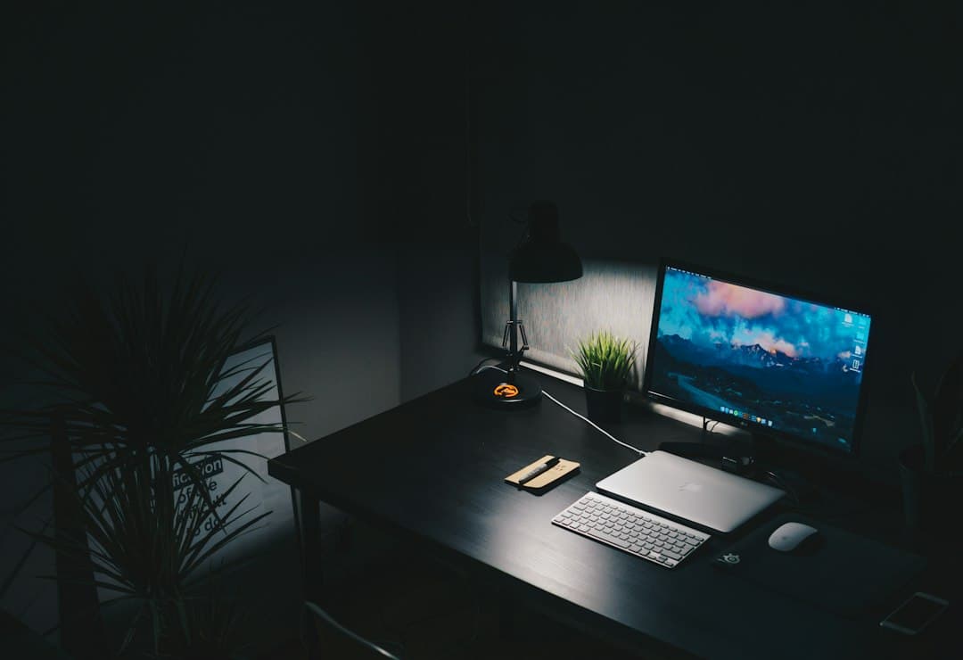 Darkly lit desk with a laptop, monitor displaying a landscape, and a plant, suggesting a faceless YouTube creator's workspace.